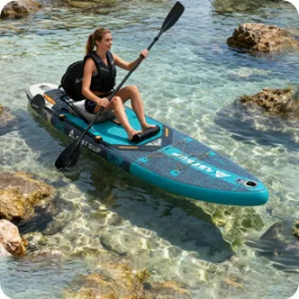 A woman in a life vest paddleboarding on a turquoise inflatable board over clear, shallow water with visible rocks.