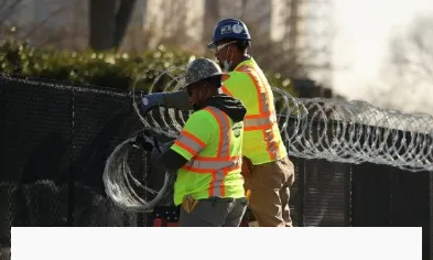 Two workers are installing cross razor wire on the top of the fence.